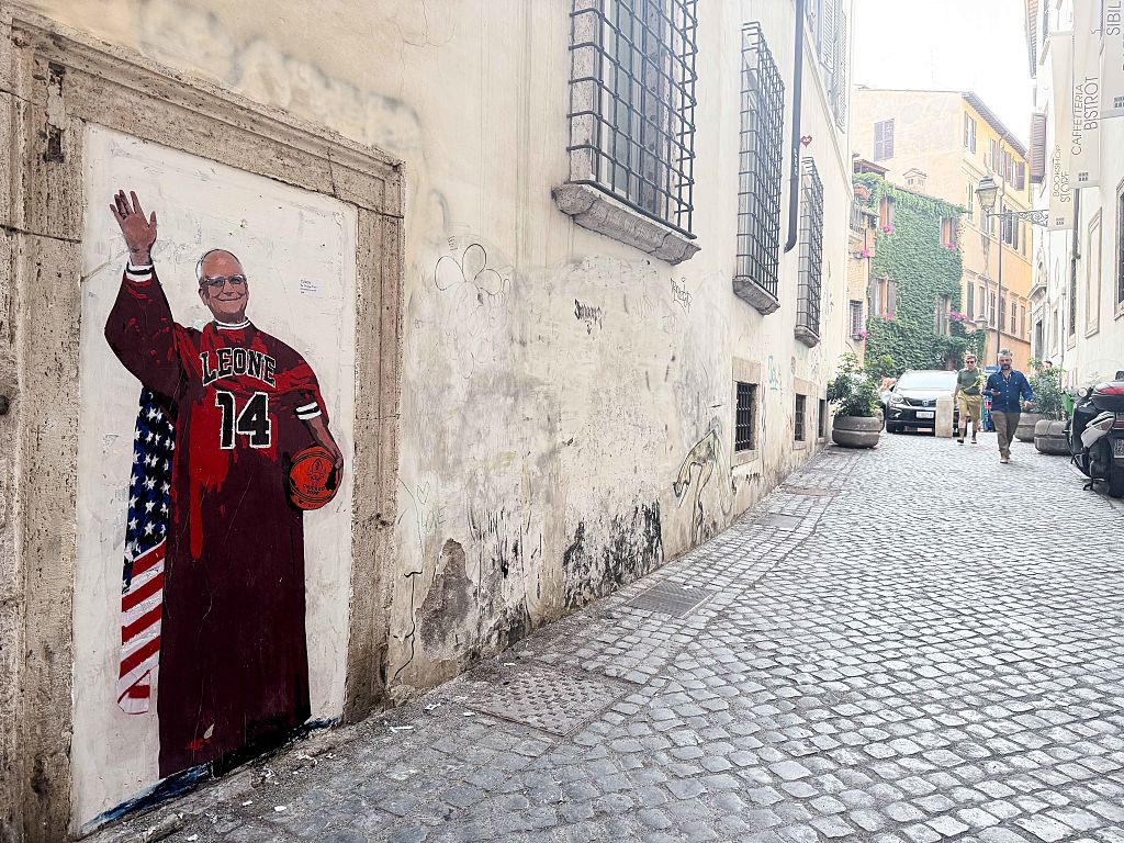 Street art of a person in a red basketball jersey holding a ball and waving, painted on a doorway along a cobblestone alley with buildings and pedestrians in the background.
