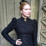 A woman wearing a high-necked, black Victorian-style dress poses near a stone wall with architectural details.