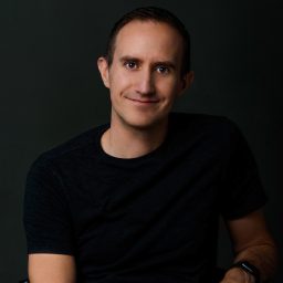 A man in a dark t-shirt sits against a dark background, smiling with his hands clasped in his lap.