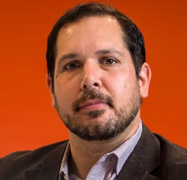 A man with short dark hair and a beard, wearing a checked shirt and dark blazer, poses in front of an orange background.