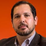 A man with short dark hair and a beard, wearing a checked shirt and dark blazer, poses in front of an orange background.