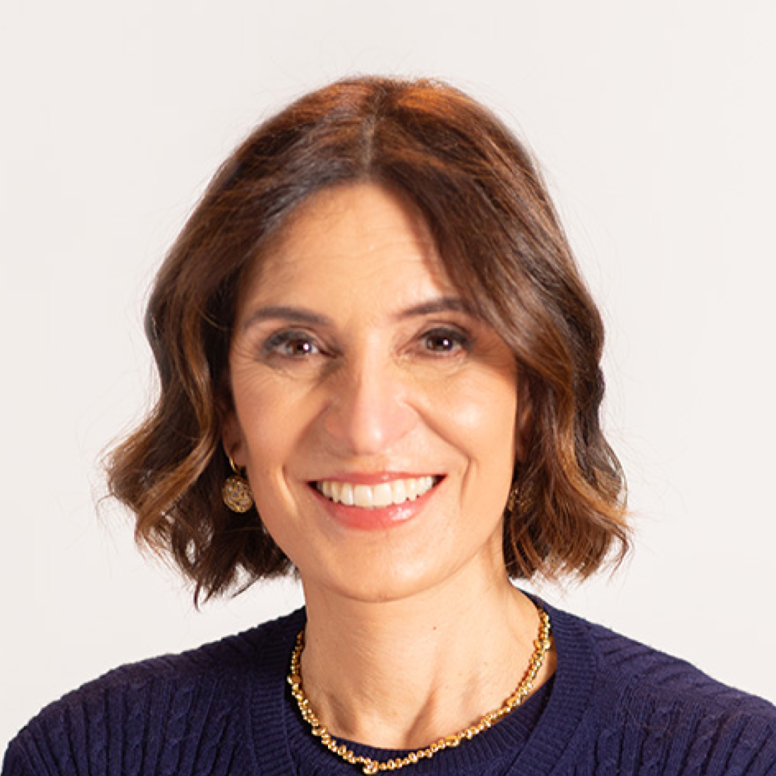 A woman with shoulder-length brown hair, wearing a navy sweater, gold necklace, and earrings, smiles at the camera against a plain light background.