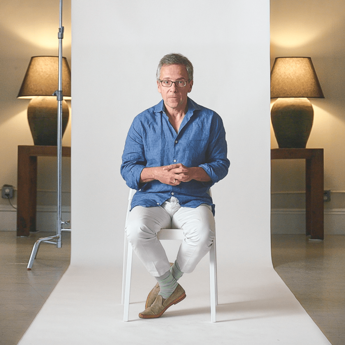 A man in a blue shirt and white pants sits on a white chair in a studio, with lamps and tables in the background and a white backdrop behind him.