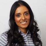 A woman with long, wavy dark hair smiles at the camera. She is wearing a gray and white striped button-up shirt and is posed in front of a plain light background.
