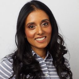 A woman with long, wavy dark hair smiles at the camera. She is wearing a gray and white striped button-up shirt and is posed in front of a plain light background.