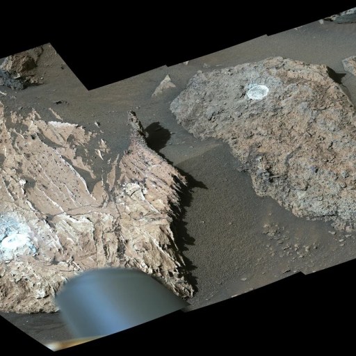 Two large Martian rocks with circular drill holes and light-colored dust are shown on a sandy, rocky surface. Part of a rover’s equipment, searching for traces of organics or signs of Mars life, is visible at the bottom of the image.