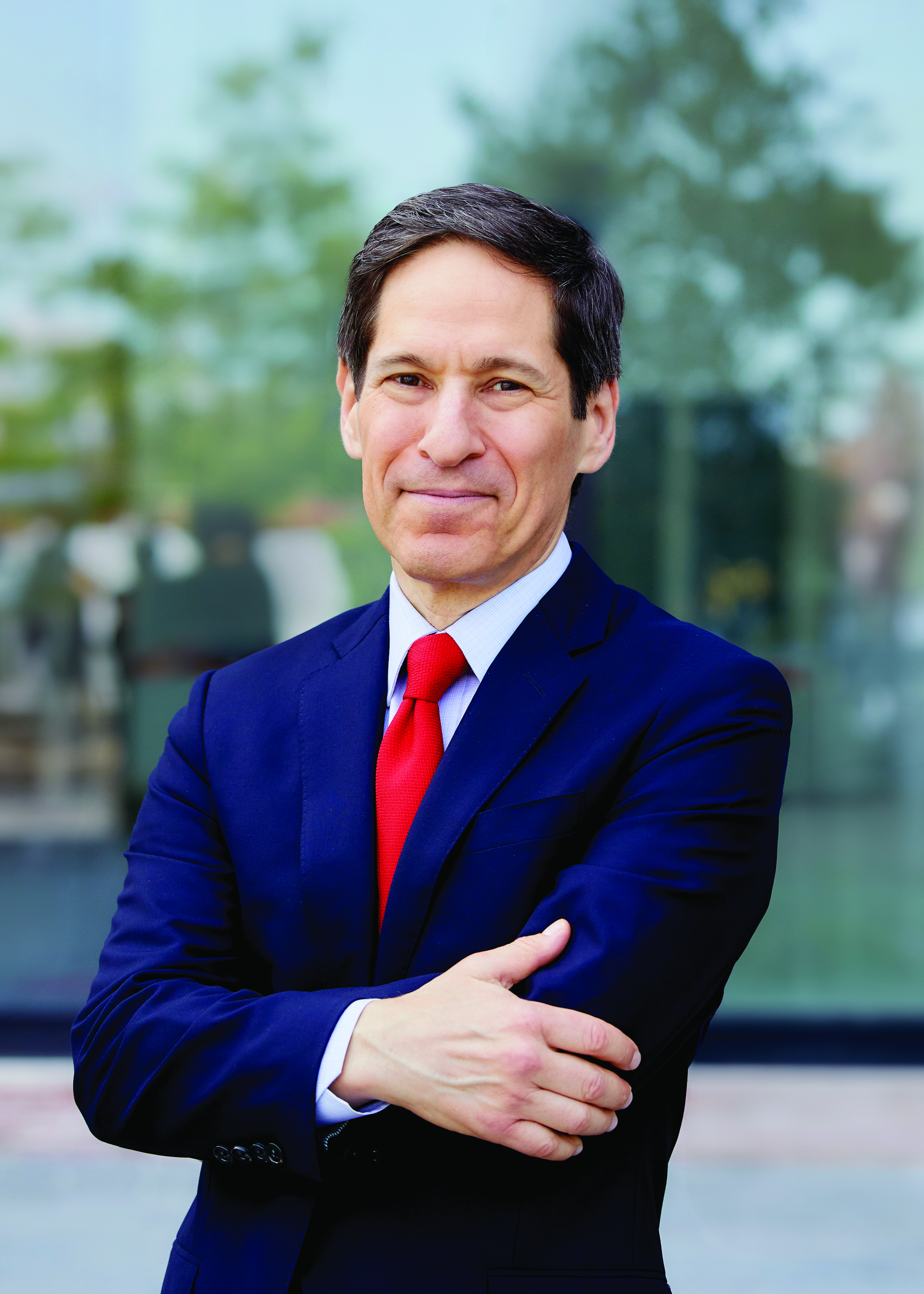 A man in a navy suit and red tie stands outdoors with arms crossed, smiling slightly at the camera.