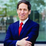 A man in a navy suit and red tie stands outdoors with arms crossed, smiling slightly at the camera.