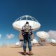 A person with a backpack stands in front of large satellite dishes in a desert area under a blue sky with scattered clouds.