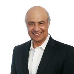 A middle-aged man with a shaved head and dark suit jacket smiles at the camera against a plain white background.