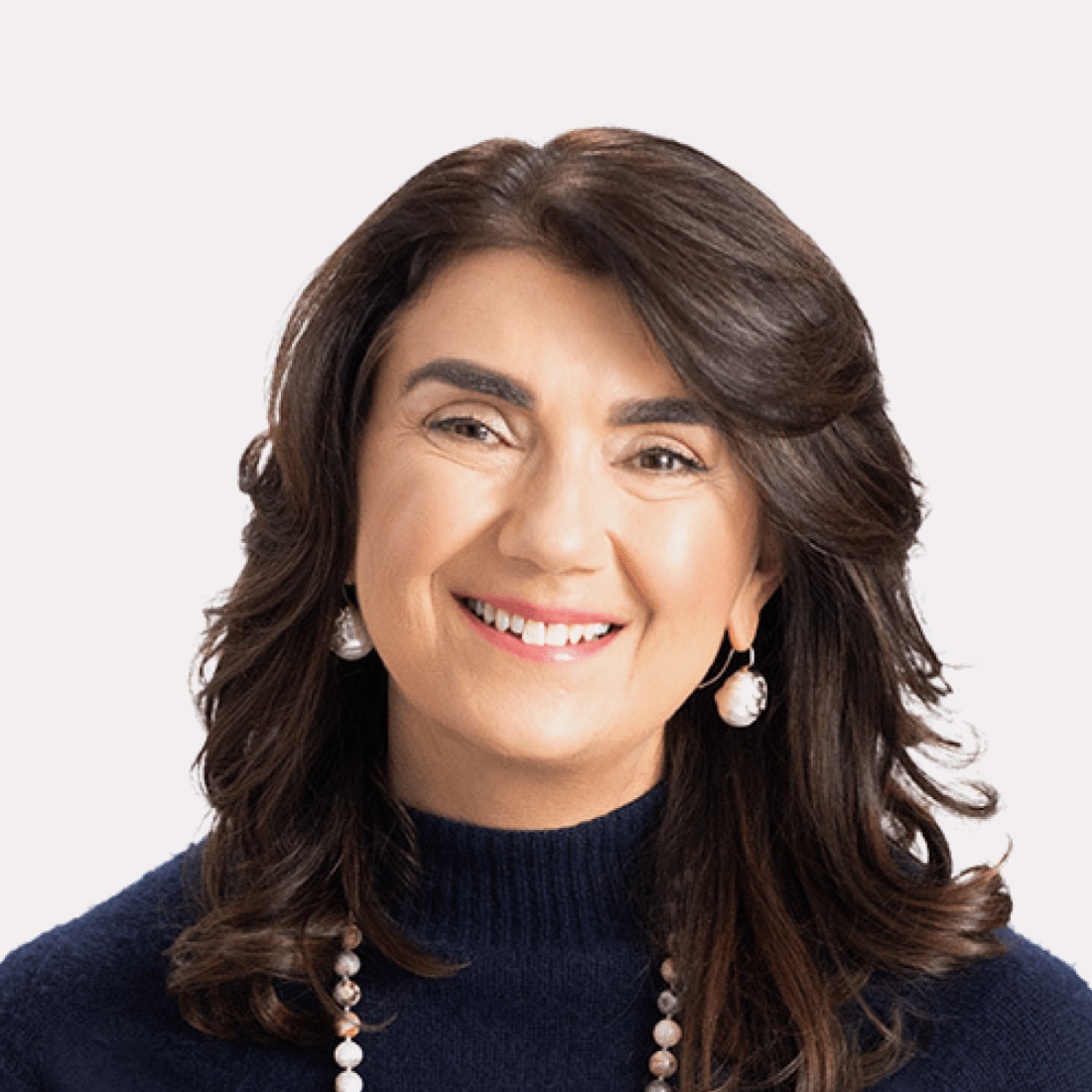 A woman with medium-length brown hair, wearing a navy sweater, pearl earrings, and a beaded necklace, smiling at the camera against a plain light background.
