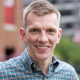 A man with short light brown hair wearing a plaid shirt smiles at the camera outdoors, with a blurred city background.