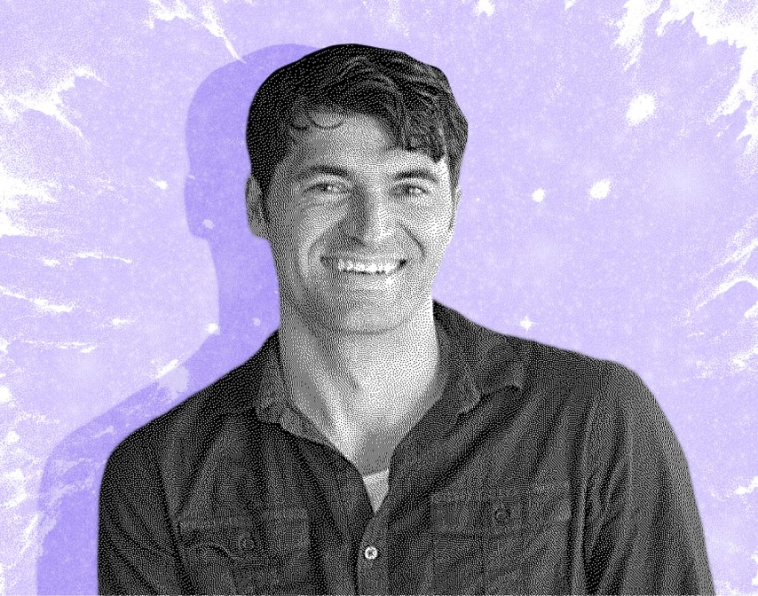 A smiling man with short dark hair wears a button-up shirt, standing in front of a purple, splattered-texture background.