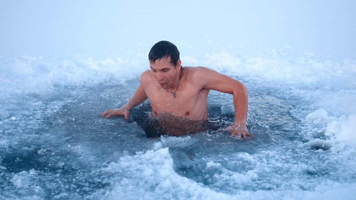 A man emerges from an ice hole in a frozen body of water, surrounded by snow and ice, taking a cold plunge during winter.
