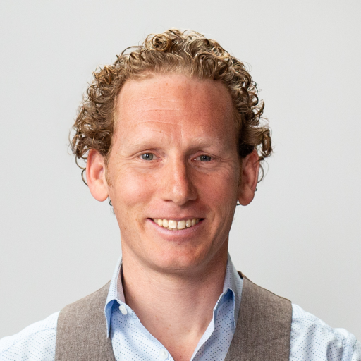 A man with short curly hair, wearing a light blue shirt and a grey vest, smiles in front of a plain light background.