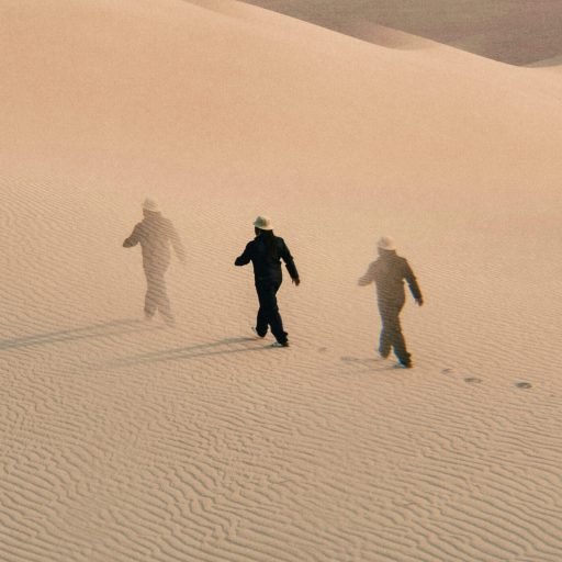 Three figures in hats walk across rippled sand dunes, leaving footprints behind them under soft, diffused light.