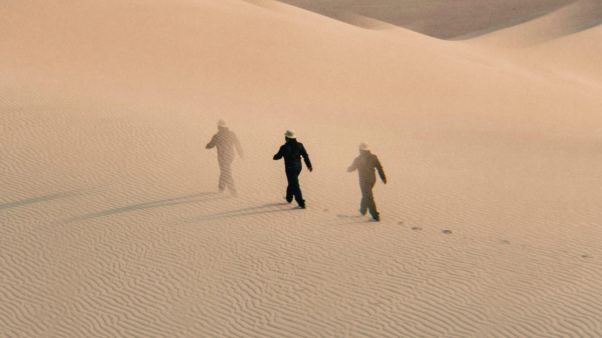 Three figures in hats walk across rippled sand dunes, leaving footprints behind them under soft, diffused light.