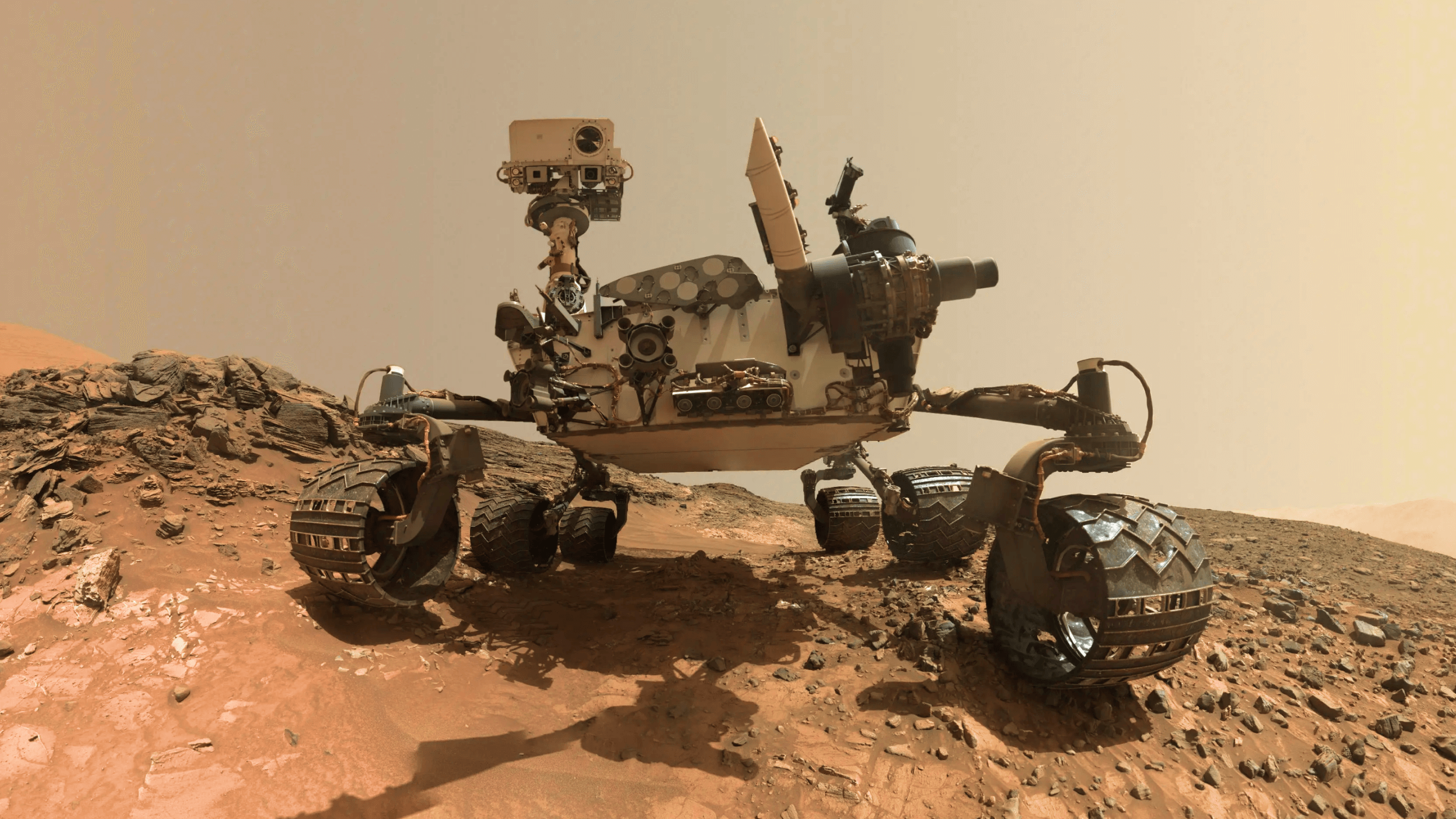 Mars rover on rocky terrain, showing its camera mast, equipment, and six wheels against a dusty, reddish Martian landscape.