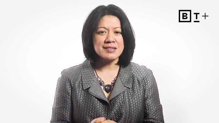 A woman with shoulder-length black hair wears a patterned gray jacket and a statement necklace, standing in front of a white background with a "B T +" logo in the top right corner.