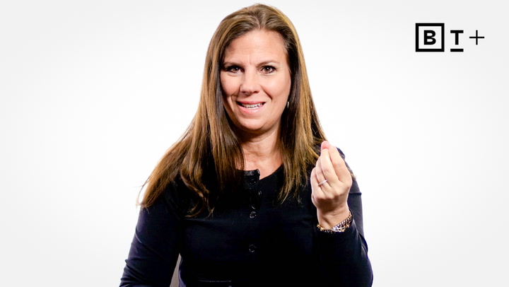 A woman with long brown hair wearing a black top gestures with her right hand against a plain white background with a "BT+" logo in the corner.