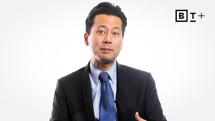 A man in a dark suit and blue tie speaks in front of a plain white background with a "BT+" logo in the top right corner.