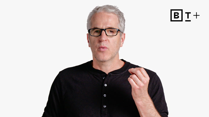 A man with gray hair and glasses wearing a black henley shirt gestures with his right hand. A "B T +" logo appears in the top right corner on a white background.