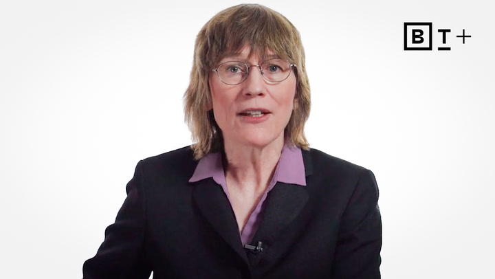 A person with shoulder-length light brown hair and glasses wearing a dark blazer and purple shirt, positioned in front of a white background with a "B T +" logo in the top right corner.