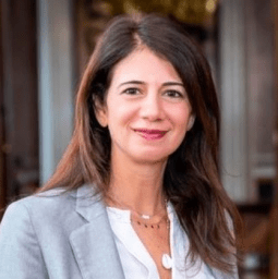 A woman with long brown hair, wearing a light gray blazer and white blouse, stands indoors and smiles at the camera.