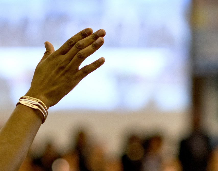 A raised hand with bracelets is shown in focus against a blurred background of people indoors.