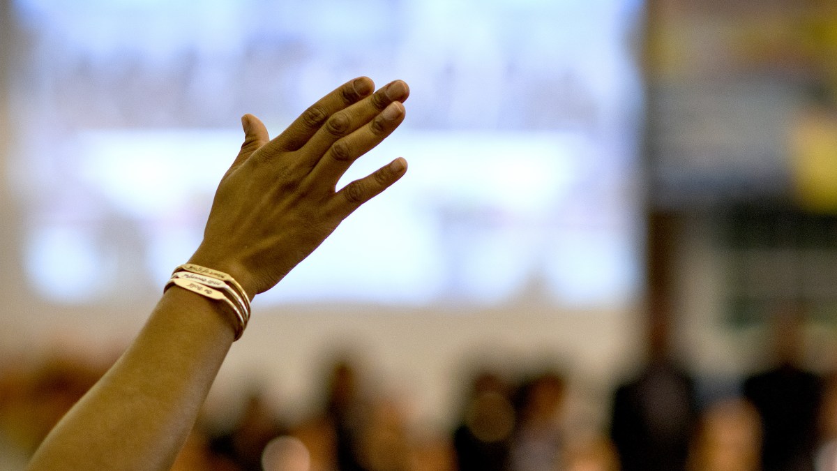 A raised hand with bracelets is shown in focus against a blurred background of people indoors.