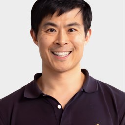 A man with short black hair smiles at the camera, wearing a dark polo shirt with a small embroidered logo on the chest, set against a plain light background.