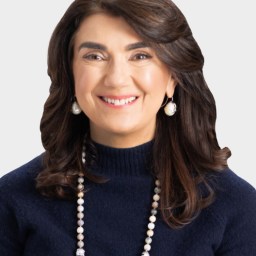Woman with shoulder-length brown hair, wearing a navy blue sweater, pearl earrings, and a beaded necklace, smiling at the camera against a plain light background.