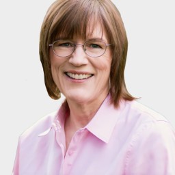 A smiling person with short, light brown hair and glasses, wearing a light pink button-up shirt against a plain background.