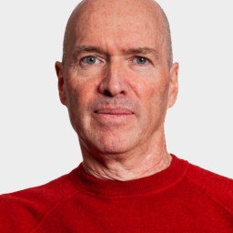 A middle-aged man with a bald head and light skin wearing a red sweater, facing forward against a plain light background.