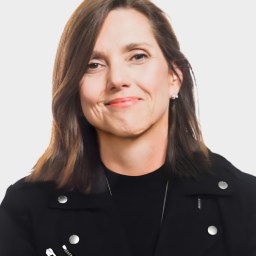 A woman with straight brown hair wearing a black jacket and a black top, smiling slightly, is posing against a plain white background.