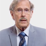 An older man with gray hair, glasses, and a mustache is wearing a light gray suit, white shirt, and striped tie, facing the camera against a plain light background.
