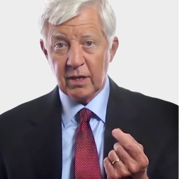 Older man with white hair in a suit and red tie gestures with his right hand while looking forward against a plain background.