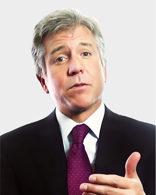 A man in a suit and polka-dot tie gestures with his right hand while speaking, against a plain light background.