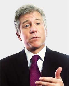 A man in a suit and polka-dot tie gestures with his right hand while speaking, against a plain light background.
