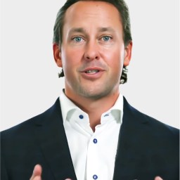 A man in a dark suit and white button-up shirt gestures with his hands while speaking, set against a plain light background.