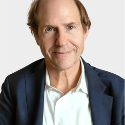 A middle-aged man with light brown hair wearing a white shirt and dark blazer, facing the camera with a neutral expression against a plain background.
