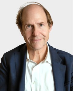 A middle-aged man with light brown hair wearing a white shirt and dark blazer, facing the camera with a neutral expression against a plain background.