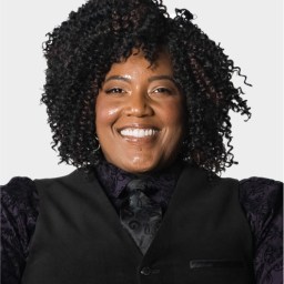 A person with curly hair, wearing a patterned dark shirt, vest, and tie, smiling in front of a plain light background.