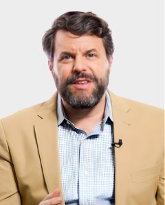 A man with a brown beard and hair, wearing a tan blazer over a checked shirt, speaks while facing the camera against a plain light background.