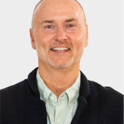 A middle-aged man with light skin, short thinning hair, and a light beard, wearing a light green shirt and a dark jacket, smiling at the camera against a plain background.