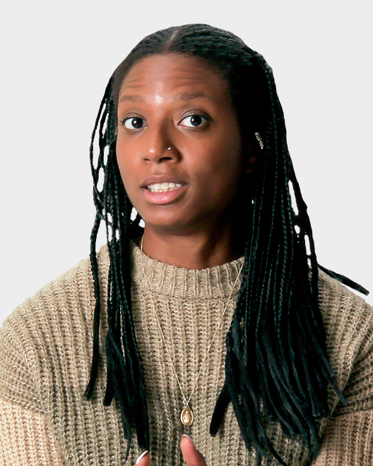 A person with long braided hair wearing a beige knit sweater and a pendant necklace, looking slightly to the side against a plain background.