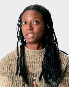 A person with long braided hair wearing a beige knit sweater and a pendant necklace, looking slightly to the side against a plain background.