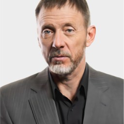 A middle-aged man with short brown hair and a beard, wearing a gray pinstripe suit jacket over a black shirt, poses against a plain light background.