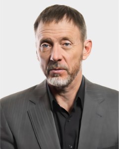 A middle-aged man with short brown hair and a beard, wearing a gray pinstripe suit jacket over a black shirt, poses against a plain light background.