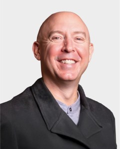 A bald man wearing a dark coat and light shirt smiles at the camera against a plain white background.